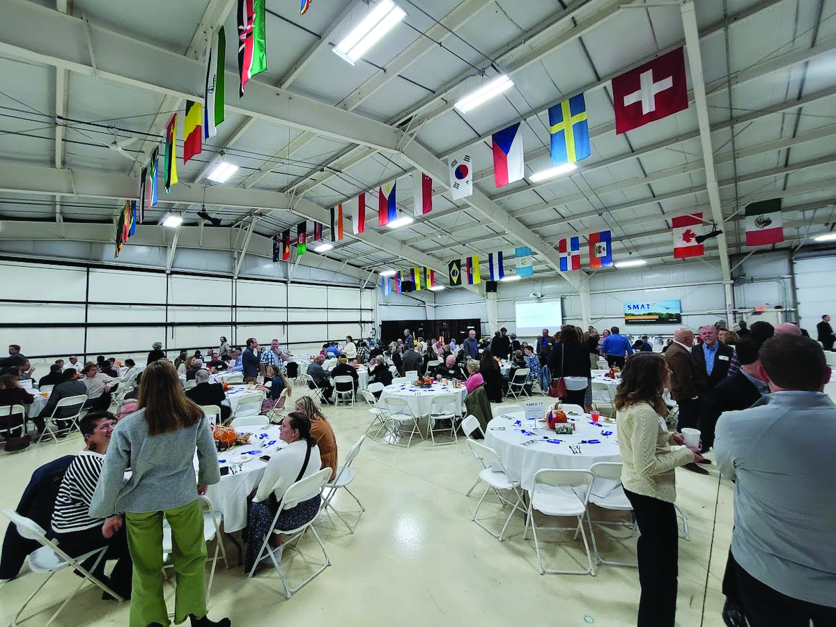 Image of people sitting at tables inside of an airplane hanger. The room is all white and there are flags hanging from the ceiling. 