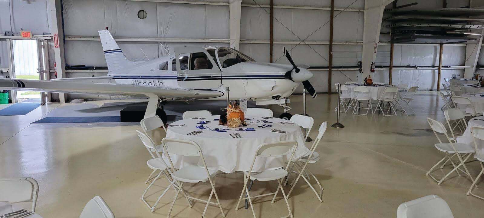 Image of tables set up for a party inside of an airplane hanger with a small airplane parked inside. 