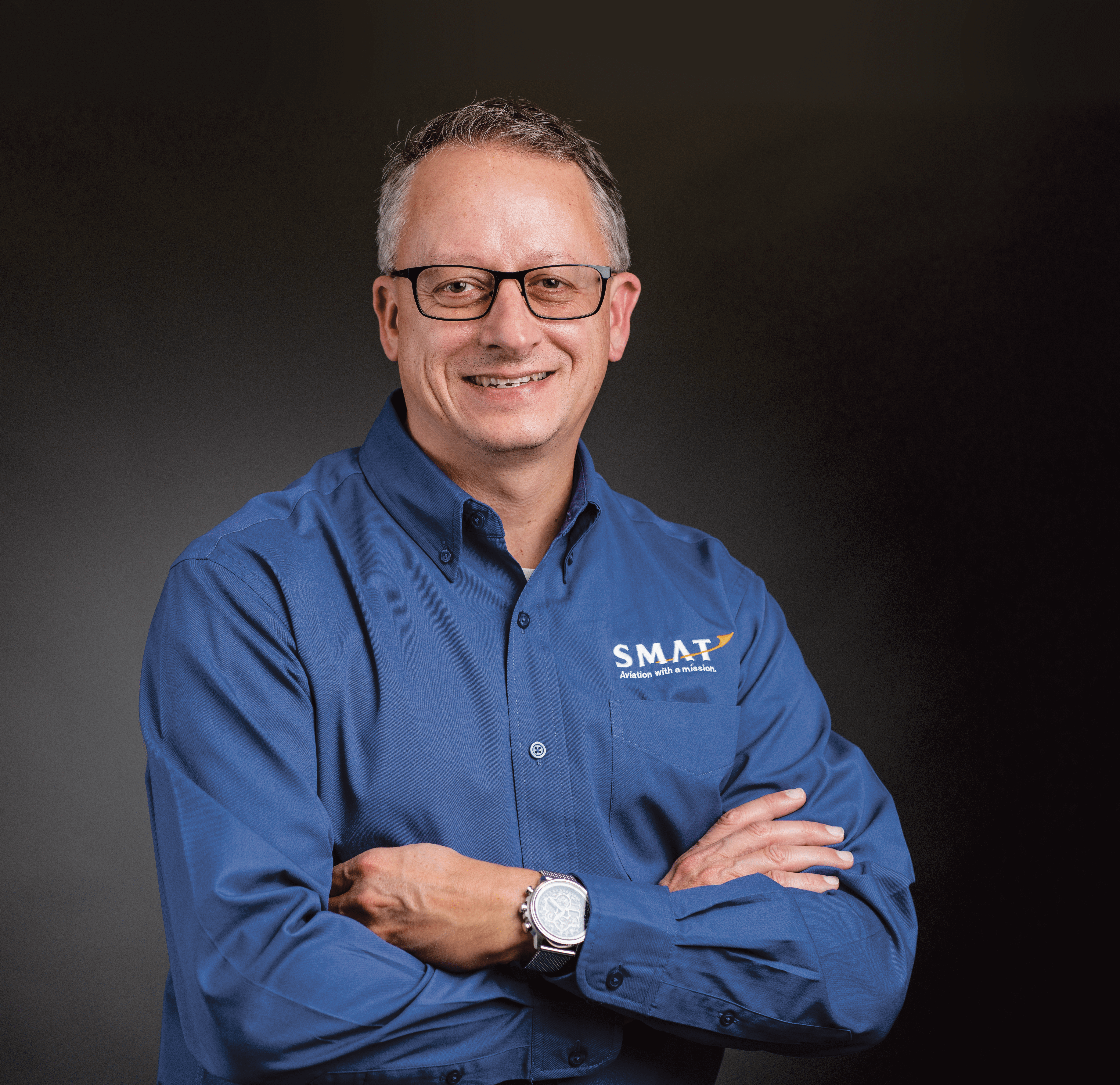 Image of the President and CEO of SMAT. He is smiling with his arms crossed in a blue button up with a dark grey background behind him. He has glasses and grey hair. 